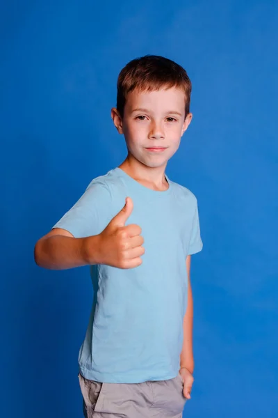 青いモックアップTシャツを着て親指で手を挙げた少年 ライトブルーのスタジオの背景の上にカメラを見ている — ストック写真