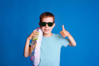 Oğlan, açık mavi stüdyo arka planında mavi tişört giyerek yaz tatilinin tadını çıkarıyor.