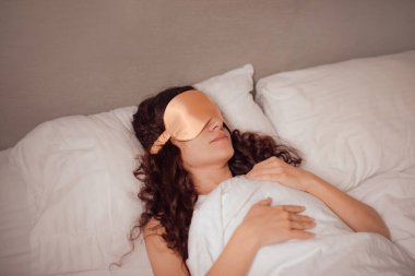 woman in beige silk sleep mask rest in bed. deeper and more restful sleep by blocking out any unwanted light clipart