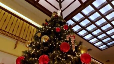 Büyük salonda kırmızı ve altın çiçeklerle süslenmiş Noel ağacı, toplar, ışıklar ve çelenkler. 