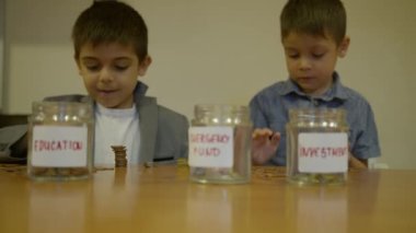 Takım elbiseli çocuk madeni paralara bakıyor ve değer başına istifliyor. Küçük kardeş parayı eğitim kavanozuna koyar. Odaklan, yavaş çekim. Yüksek kalite 4k görüntü