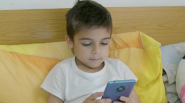Tatlı beyaz çocuk yatakta uzanıyor ve telefonda çizgi film seyrediyor. Yüksek kalite 4k görüntü