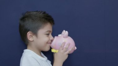 Çocuk tasarrufu konsepti. Kafkasyalı çocuk komik suratlı domuz kumbarasıyla oynuyor. Mavi arkaplanda kopyalama alanı ile izole edilmiş. Ağır çekim. Yüksek kalite 4k görüntü