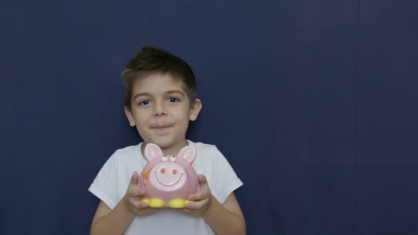 儿童储蓄概念 白人男孩用滑稽的面部表情摇动着小猪银行 在蓝色背景上与复制空间隔离 慢动作高质量的4K镜头 — 图库视频影像