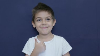 Sevimli küçük çocuk baş parmağını kaldırıyor, mutlu yüz ifadesi, duygu, mavi arka planda yavaş çekim. Yüksek kalite 4k görüntü