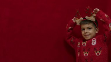 Ren geyiği kostümlü şirin bir çocuk ve kamera önünde kafa bandı kulakları utangaç oluyor. Kırmızı arka planda izole edilmiş. Ağır çekim. Yüksek kalite 4k görüntü