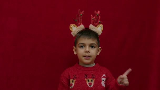 クリスマスのセーターでかわいい男の子とトナカイのアントラーは 赤い背景で隔離された空のスペースに指を指します スローモーション 高品質の4K映像 — ストック動画