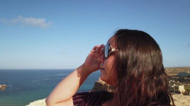 Genç bir kadın, Malta 'nın Tuffieha Körfezi manzaralı tepedeki güneş gözlüğü ve saçlarını düzeltiyor. Yüksek kalite 4k görüntü
