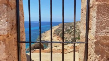 Comino Adası 'ndaki Santa Maria Bataryası. Camdaki metal parmaklıklardan ay plajına ve kayalıklara doğru bakıyor. Yüksek kalite 4k görüntü