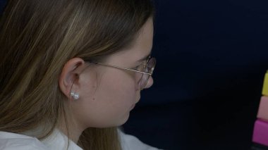 Bir kadın çalışma alanında oturur, işine derinden konsantre olur, dikkatini ve dikkatini gösterir. Gözlük takıyor ve çevresi verimlilik ve adanmışlığı vurgulayan profesyonel bir düzenekle çevrili.