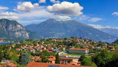 İtalya, Como Gölü 'ndeki Bellagio köyü. Dağların ve yeşil ormanların arasında park ve antika şehirlerin olduğu tepede panoramik manzara. Su, dalgalar ve bulutlu mavi gökyüzü ile yaz resimli manzara.