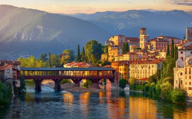 Bassano del Grappa, Veneto, Italy. Bridge Ponte degli Alpini at river Brenta. Panoramic view old town with vintage building tower, wooden Alpine mountains scenic sunset landscape