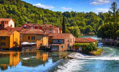 Old village Borghetto Valeggio sul Mincio, Veneto region, Italy. Panoramic view at the town houses on river. Green forest and blue sky. Sunny day