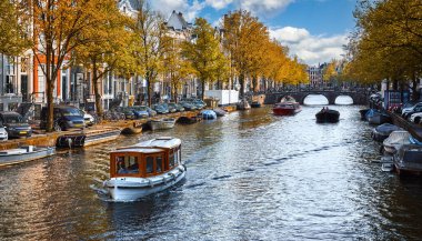 Amsterdam Hollanda 'da Amstel nehrinin altındaki evlerde. Eski Avrupa kentinin günışığıyla dolu olan son bahar manzarası. Suyun üstünde turistlere eğlence tekneleri