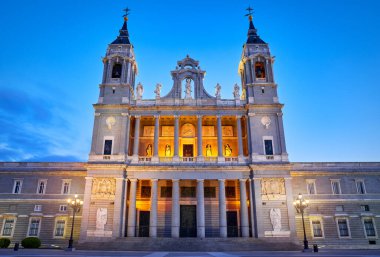 Madrid, İspanya. Santa Maria la Real de la Almudena Katedrali 'nin Plaza de la Armeria' daki ön manzarası. Günbatımı güneşi, çiçekler ve yeşil çalılarla ünlü bir yer. Işıklı sokak lambaları ve mavi gökyüzü.