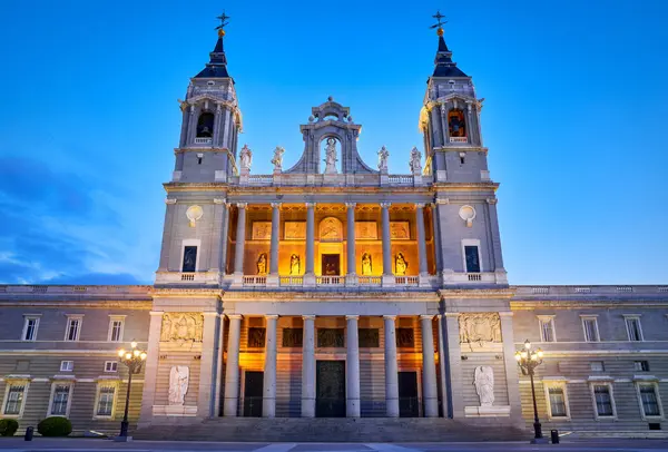 Madrid, İspanya. Santa Maria la Real de la Almudena Katedrali 'nin Plaza de la Armeria' daki ön manzarası. Günbatımı güneşi, çiçekler ve yeşil çalılarla ünlü bir yer. Işıklı sokak lambaları ve mavi gökyüzü.