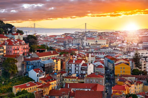 Lizbon, Portekiz. Günbatımının en güzel manzarası. Alfama bölgesinin tarihi eski kasabasındaki evlerde akşam manzarası. 25 Nisan Köprüsü uzakta. Gökyüzü dramatik bulutlarla ve güneşle.