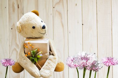 Kahverengi ayıcığın elinde hediye kutusu var. Özel bir gün. Arka planda çiçek aranjmanıyla mutlu.