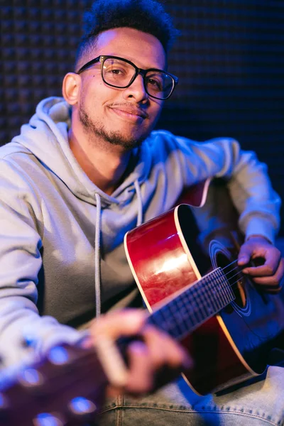 stock image Young afro musician wearing glasses playing acoustic guitar, on dark background