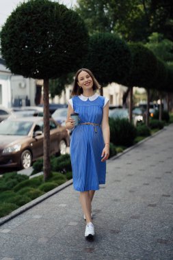 Mavi elbiseli güzel hamile bir kadın şehir sokaklarında yürüyor ve içki içiyor..