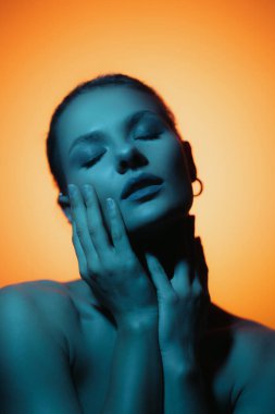 Fashion woman profile portrait on orange gradient background with soft blue light play on skin. Hands touching skin. Lights play.