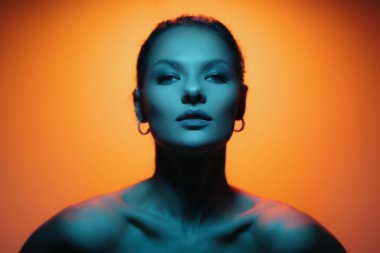 Fashion woman profile portrait on orange gradient background with soft blue light play on skin. Model looking at camera. Lights play.