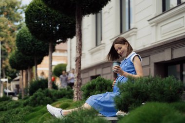 Mavi elbiseli hamile bir kadın şehirde oturup dinlenirken karnına dokunuyor.