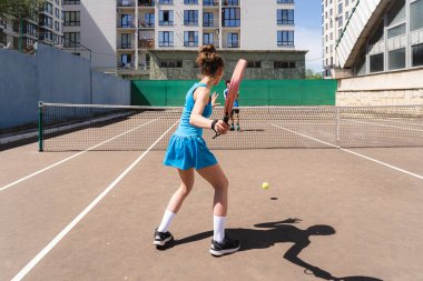 Padel raketle tenis oynayan tanınmayan genç bir kızın gölgeyle sahada dikilmesi ve çevresi binalarla çevrili isimsiz bir çocukla oynaması.