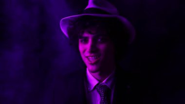 Young curly haired brunette man with hat and suit looks at the camera and smiles