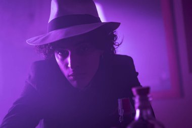 A young man with curly, brunette hair is looking at the camera with a smile. He is wearing a hat and suit. He appears confident and poised. The dark background adds a sense of mystery and intrigue.