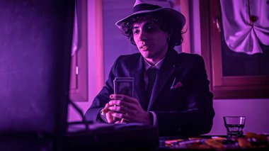 A drunk young man in a black suit and hat is celebrating his win at gambling. He is putting his money into a briefcase placed in front of him at the poker table.