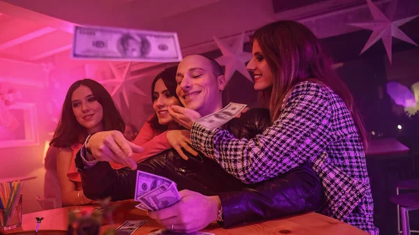 A man with a stack of cash and surrounded by women. Throws away some of the cash on the table. He is likely gambling. Suggests a concept of luxurious nightlife, gambling, and entertainment.