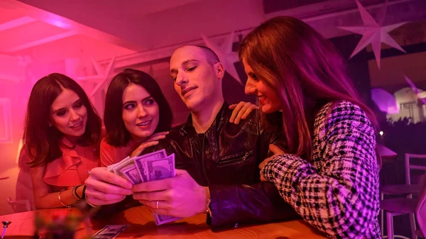A man with a stack of cash and surrounded by women. Throws away some of the cash on the table. He is likely gambling. Suggests a concept of luxurious nightlife, gambling, and entertainment.