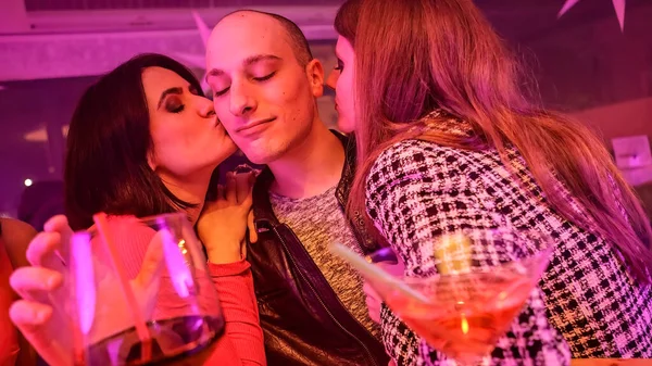 A man at a bar with a group of women flirtatiously interacting with him. Some are touching him and one woman is leaning in to give him a kiss. The man is holding a wine glass and enjoying the attention.