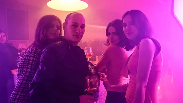 A man at a bar with a group of women flirtatiously interacting with him. Some are touching him and one woman is leaning in to give him a kiss. The man is holding a wine glass and enjoying the attention.