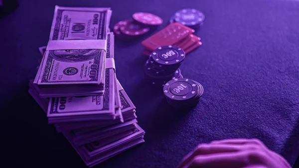 Stack of poker chips in a range of colors and a wad of dollars on the gaming table in a dimly lit casino. The chips and money suggest a game of poker is about to begin.