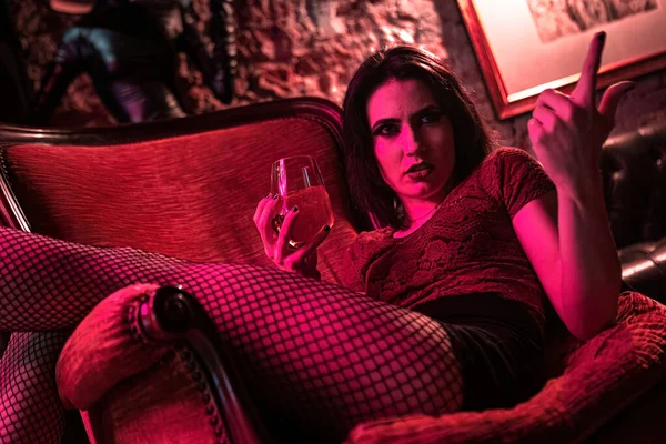 This powerful photo captures an angry brunette girl sitting on an elegant vintage armchair, holding a drink and glaring intensely. Great for illustrating emotions and storytelling in various projects.
