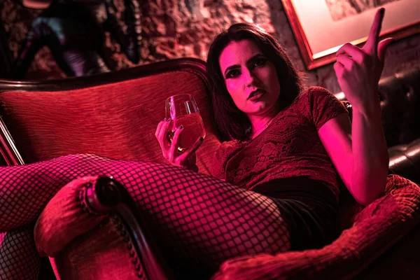 This powerful photo captures an angry brunette girl sitting on an elegant vintage armchair, holding a drink and glaring intensely. Great for illustrating emotions and storytelling in various projects.