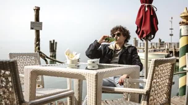 2023年4月11日 意大利锡尔敏 一个孤独的年轻人坐在码头上的酒吧桌旁 一边喝啤酒 一边忧郁地看着自己 — 图库视频影像