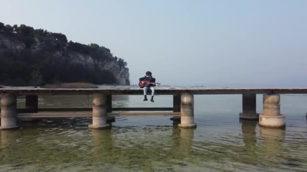 Ein Einsamer Junge Spielt Gitarre Während Auf Einer Seebrücke Sitzt — Stockvideo