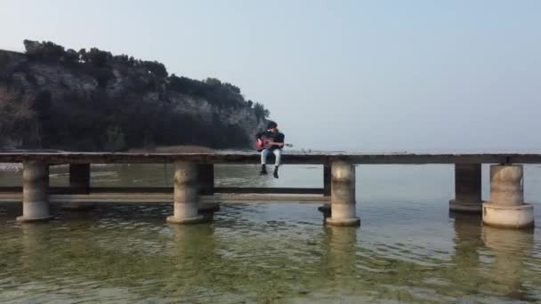 Ein Einsamer Junge Spielt Gitarre Während Auf Einer Seebrücke Sitzt — Stockvideo