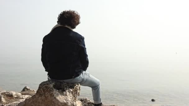一个年轻人孤零零地坐着 面色忧郁 凝视着一个雾蒙蒙的湖面 雾气从湖面升起 — 图库视频影像