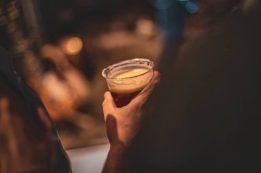 Plastik bardakta bira tutan bir elin yakın plan fotoğrafı. Arka planı bulanık..