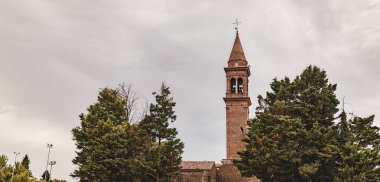 Tuğla yüzlü çan kulesi, bulutlu bir arka plana karşı ağaç tepelerinin üzerinde yükseliyor.