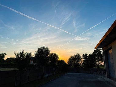 Kimyasal yollar, gün batımında gökyüzünde ilerleyen kontrastlar, ön planda ağaç ve bina siluetleri.