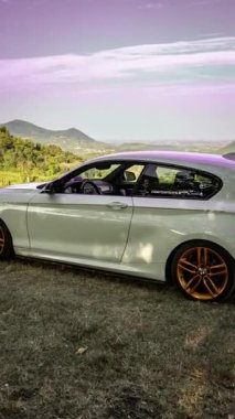Calaone, İtalya 28 Ağustos 2024: Açık bir gökyüzü altında manzaralı tepeler ve ağaçlarla dolu çimenli bir alana park edilmiş BMW 1 Serisi F21 yan görüntüsü.