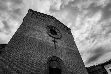 Karanlık bir gökyüzünün altında haç ve dairesel pencereli tuğla kilise cephesinin siyah-beyaz perspektifi.