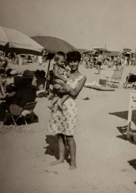 Roma, İtalya 1964 olabilir: Çocuğunu kalabalık bir plajda kucaklayan bir annenin yaz gününün keyfini çıkaran diğer aileler tarafından çekilmiş klasik siyah beyaz fotoğrafı