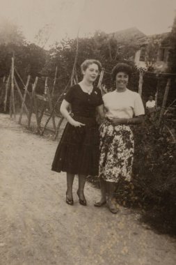 Roma, İtalya 1964 olabilir: İki zarif giyimli kadın, ahşap bir çitin yakınındaki toprak bir yolda birlikte poz vererek, 20. yüzyılın ortalarında kırsal hayatın bir anını yakalıyorlar.