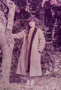 Roma, İtalya 1964 olabilir: Bahçedeki bir ağaca yaslanmış, süet ceket giymiş, 1970 'lerin modasını yansıtan kıvırcık saçlı, gülümseyen genç kadın